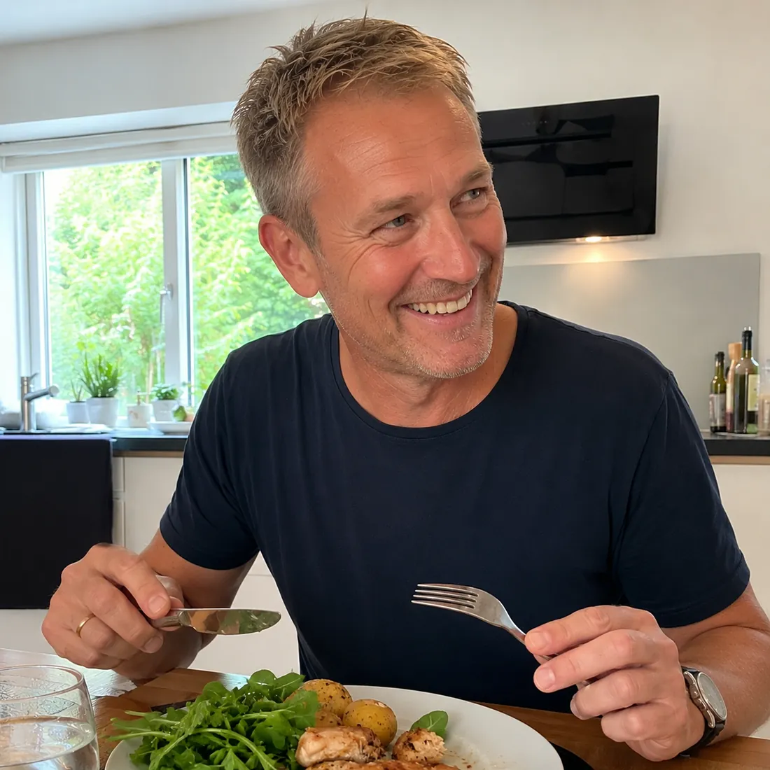 Mand smiler og spiser en tallerken med kylling, kartofler og salat i sit køkken